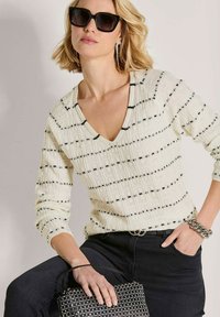 Frau trägt schwarze Sonnenbrille, weißen V-Ausschnitt-Pullover mit schwarzen Streifen, schwarze Hose, hält eine schwarze Nietenhandtasche und ein silbernes Armband.