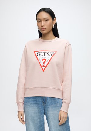 Ung kvinde med langt sort hår iført en lyserød GUESS-sweatshirt med rød trekantlogo og blå jeans, stående mod en ensfarvet hvid baggrund.