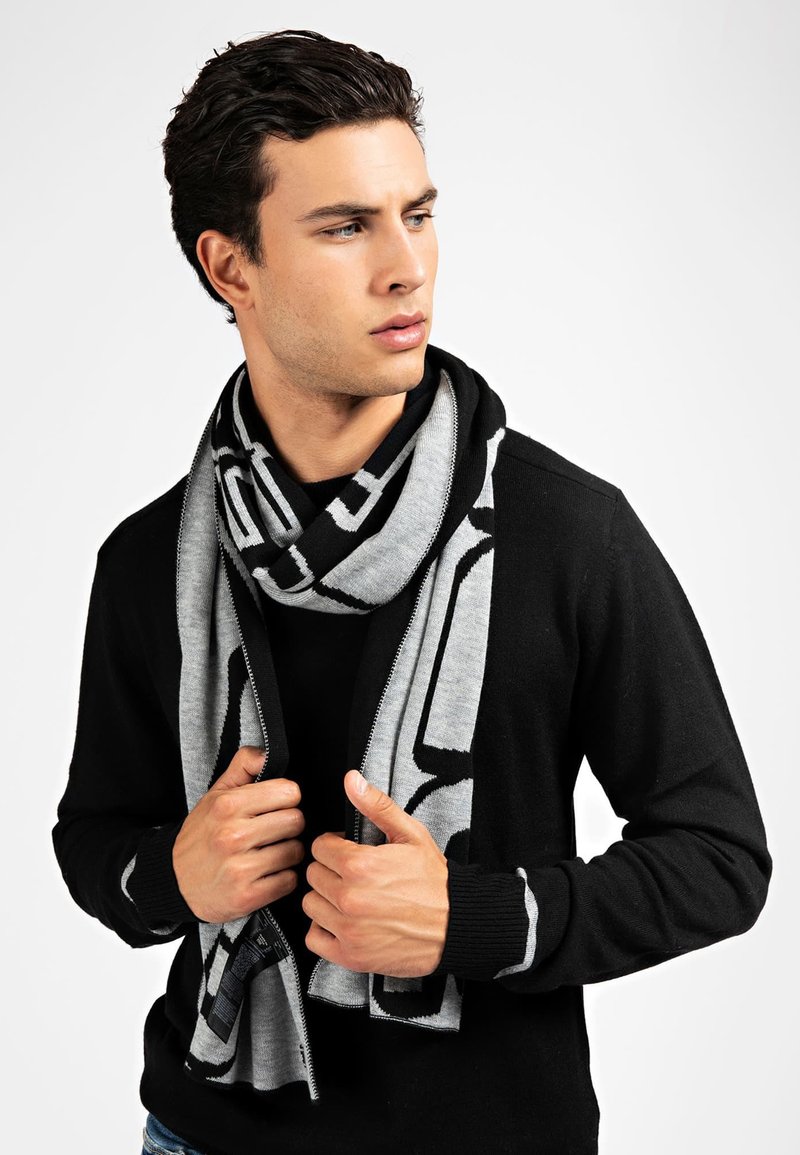 Écharpe grise et noire à motif drapée autour du cou ; texture tricotée. Portée avec un pull noir zippé. Gros plan de mains tenant l'écharpe.