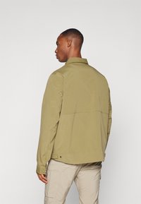 Veste vert olive clair, fabriquée en tissu lisse, dotée d'un col et de poignets boutonnés, avec un design arrière épuré et un léger détail d'ouverture.