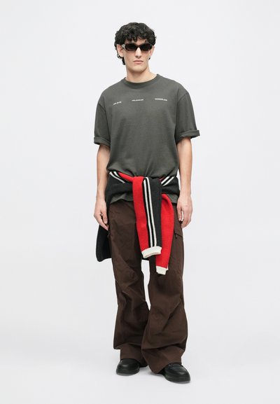 Siv bombažna majica s povitimi rokavi, rjave široke hlače in pleten pulover zavezan okoli pasu, ki ima rdeče, črne in bele stripe.