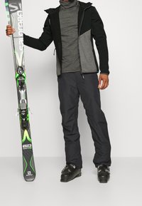 Ski outfit featuring a black and grey zip-up jacket, grey turtleneck, black ski pants, and black ski boots. Holding green-accented skis.