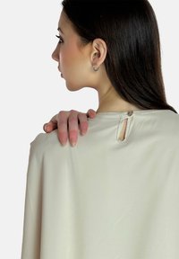 Blusa beige con una texture liscia, dotata di un'apertura a forma di chiave e bottone sul retro. Maniche lunghe e fit rilassato.