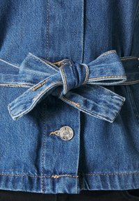 Jeanová bunda s uzlovým detailem v pase. Vybavena stříbrnými patentními knoflíky a viditelným kontrastním stehem. Tmavě modrá barva s texturovaným povrchem.