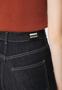 Jeans de ganga azul escuro com costura amarela, apresentando um rótulo preto na parte de trás com o logótipo "DRDENIM" e um detalhe no bolso traseiro.