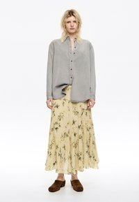 Grijze button-up shirt met lange mouwen, gecombineerd met een gele bloemenprint plooienrok en een paar bruine suède instappers.
