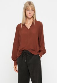 Roestkleurige blouse met een v-hals, lange mouwen en een ontspannen pasvorm, gecombineerd met zwarte broek met trekkoord en een ribbelstructuur.