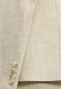 Gros plan sur la manche d'une veste en tissu beige texturé avec quatre boutons ronds assortis en rangée verticale près du poignet.