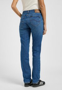 Jeans de mezclilla azul con corte recto, que presentan una etiqueta de cuero marrón de marca en la cinturilla y costuras sutiles en los bolsillos traseros.