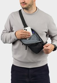 Černá, kompaktní sling taška se zipem, s šedým vnitřkem. Nastavitelný popruh. Uživatel vytahuje chytrý telefon z přední kapsy.
