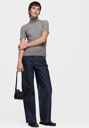 Mujer vestida con un suéter gris de manga corta y cuello alto, vaqueros anchos oscuros y zapatos negros sin cordones, sosteniendo un pequeño bolso negro a su lado.