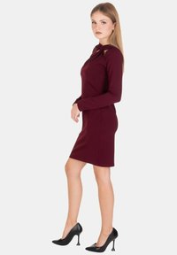 Abito bordeaux a maniche lunghe con silhouette aderente, caratterizzato da un dettaglio a nodo al collo e una texture liscia, abbinato a scarpe nere con tacco alto.