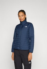 Veste isolée marine avec texture matelassée, col montant, fermeture éclair avant et poches latérales. Dispose d'un logo sur la poitrine et de poignets ajustables.