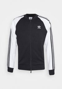 Černobílá mikina na zip s raglánovými rukávy, třemi bílými pruhy na každém rukávu a malým černým logem na hrudi.