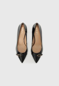 Zapatos de tacón alto de charol negro con punta afilada, que presentan un pequeño detalle de hebilla dorada y una plantilla perforada beige.