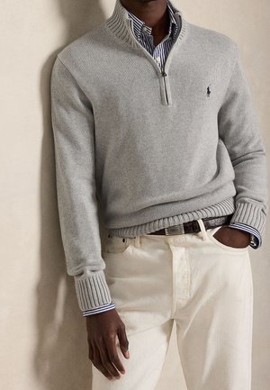 Mann trägt hellgrauen Reißverschluss-Pullover über blau gestreiftem Hemd, weiße Hose und dunklen Gürtel, mit einer Hand in der Tasche und lehnt an einer einfarbigen Wand.