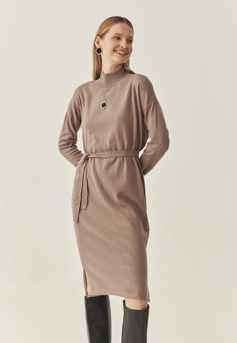 Neulemekko midi-pituudessa, savunharmaa, korkea kaulus, vyötäröllä vyö, sivuhalkiot ja pitkät hihat, yhdistetty mustiin polven korkeisiin saappaisiin.