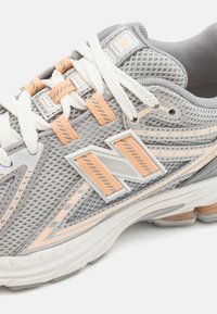 Γκρι αθλητικό παπούτσι κατασκευασμένο από διχτυωτό υλικό με πορτοκαλί λεπτομέρειες. Περιλαμβάνει λευκές κορδόνες, μια έντονη λογότυπο και μια μαξιλαρωμένη σόλα για στήριξη.