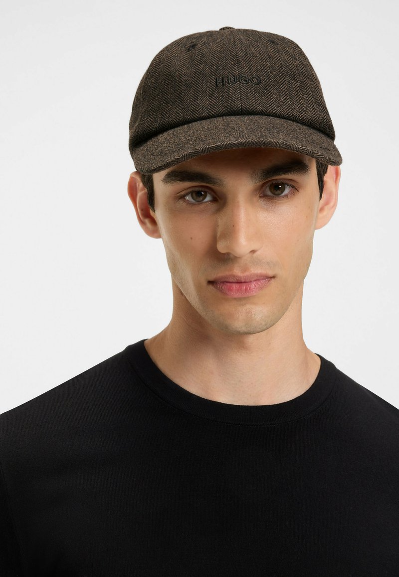 Brown herringbone cap with a rounded peak and tonal embroidered logo. Worn by a person in a black shirt against a plain background.
