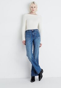 Maglione bianco a coste leggero con maniche lunghe, abbinato a jeans in denim blu a vita alta con dettagli sulle tasche e gambe leggermente svasate.