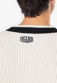 Knitted cream sweater with black ribbed collar; features vertical texture and embroidered "PEGADOR" logo at the back.