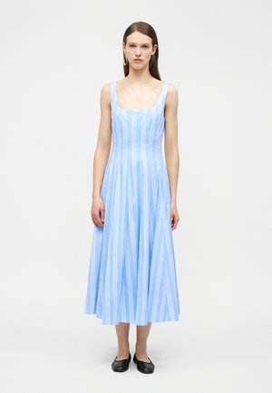 Kobieta w bezrękawną, jasnoniebieską sukienkę midi w białe pionowe paski i czarne baleriny, stojąca na jednolitym białym tle.