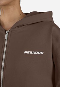 Hnedá mikina na zips vyrobená z textúrovanej látky, s predným logom "PEGADOR" v bielej farbe. Obsahuje veľkú kapucňu a zips po celej dĺžke.