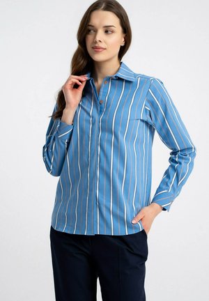 Kobieta ubrana w niebieską koszulę z długim rękawem w białe i czarne pionowe paski, pozująca z jedną ręką przy kołnierzu, a drugą w kieszeni.