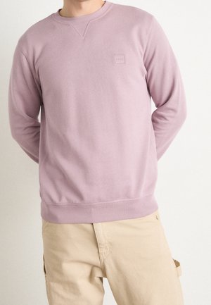 Hombre con sudadera de cuello redondo color rosa pastel y pantalones beige, de pie con las manos detrás de la espalda frente a un fondo liso.