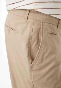 Beige Baumwollhosen mit Gürtelschlaufen, Seitentaschen und einer mit Knopf verschlossenen Gesäßtasche, getragen zu einem weiß-braun gestreiften Hemd.