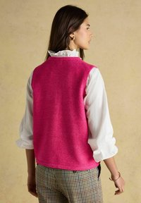 Hellrosa ärmellose Weste aus weichem Material, getragen über einem weißen, mit Rüschen versehenen Hemd, kombiniert mit karierten Hosen. Einfaches Design, keine sichtbaren Muster.