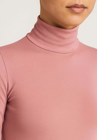 Maglia a collo alto aderente in tessuto morbido ed elastico. Color rosa chiaro con una texture liscia e cuciture minime, che esaltano una silhouette slanciata.