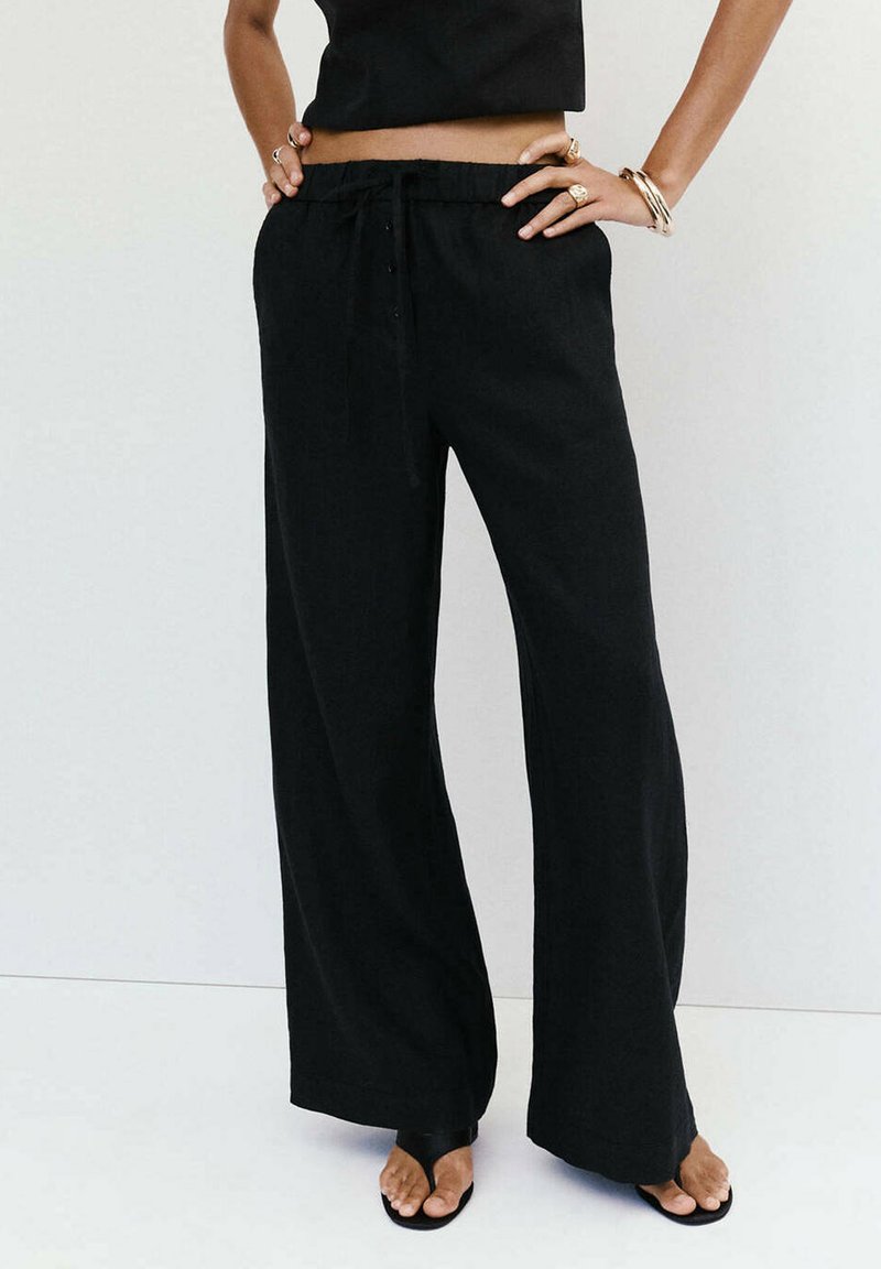 Personne portant un pantalon noir ample, un crop top noir, des bagues et bracelets en or, debout les mains sur les hanches sur fond blanc.