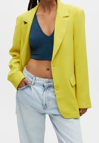 Personne portant un blazer jaune vif sur un haut court bleu foncé, avec un jean taille haute bleu clair, une main dans la poche.