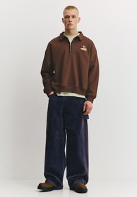 Brun fleece-pullover med en halv lynlås og lille logo, kombineret med løstsiddende mørke denimjeans og brune sko. Enkel, afslappet stil.