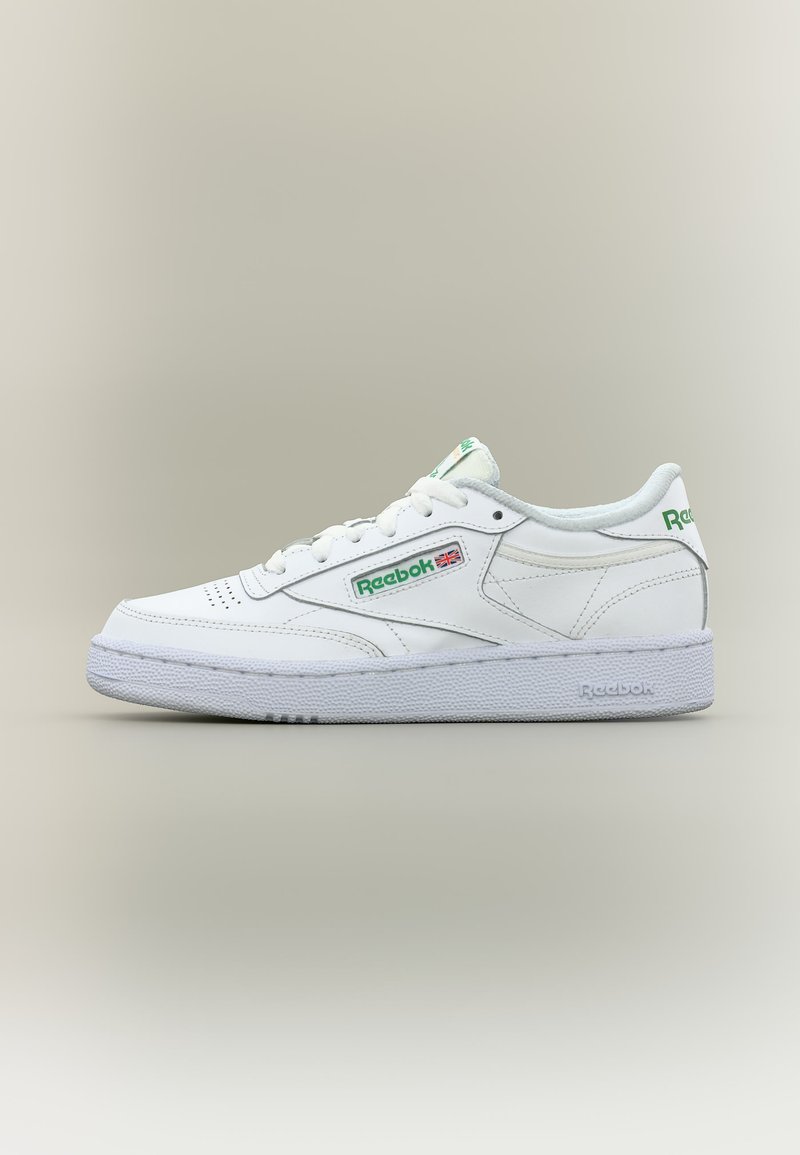 Fehér, alacsony szárú Reebok sneaker zöld logóval és az Egyesült Királyság zászlajával az oldalon, fehér talppal és fűzőkkel, semleges háttéren kívülről megjelenítve.