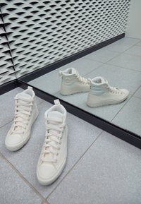Baskets montantes blanches en tissu texturé, avec des lacets et un logo circulaire sur un côté. Reflétées dans un miroir à côté.