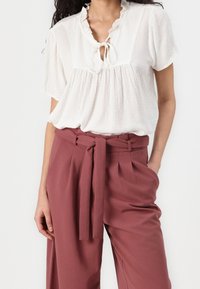 Blouse blanche texturée avec un col à nouer et un col volé, associée à un pantalon taille haute couleur rouille avec une ceinture et des plis.