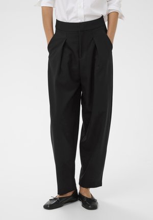 Mustad kõrge vöökohaga mustad püksid, millel on ees voldid ja küljetaskud, omades siledat tekstuuri ja vabalt voolavat lõiget, koos siledate mustade kingadega.