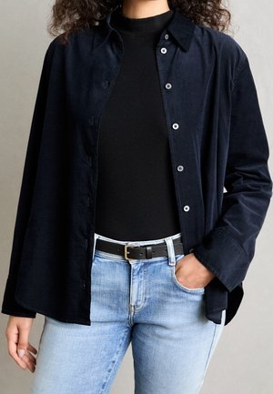 Person wearing unbuttoned dark navy shirt over black ribbed top, light blue jeans, and black belt with silver buckle, hand in pocket.