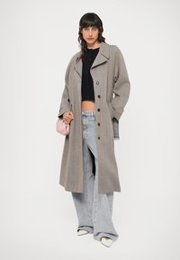 Grijze lange jas met een ingesneden kraag, knoopsluiting en een losse pasvorm, gedragen over een zwarte crop top en lichtblauwe wijde jeans.