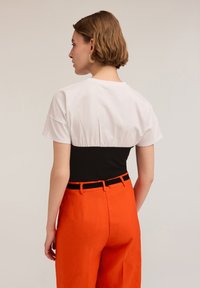 Camisa bicolor en blanco y negro con cintura fruncida y mangas cortas, combinada con pantalones naranjas de cintura alta que presentan un cinturón negro.