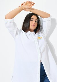 Camicia bianca a maniche lunghe con bottoni, realizzata in tessuto testurizzato. Presenta un design ricamato sul petto e una vestibilità comoda.