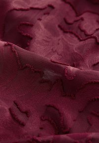 Gros plan sur un tissu bordeaux avec des motifs floraux en relief, texturés et des plis doux.