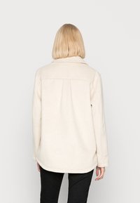 Kremowa polarowa kurtka z kołnierzem, detalem w formie pliski na plecach oraz zaokrąglonym dołem. Materiał wydaje się miękki i teksturowany.