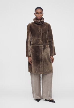 Donna che indossa un cappotto di pelliccia marrone con collo alto, pantaloni beige a gamba larga e ballerine nere con punta appuntita, in piedi contro uno sfondo neutro.