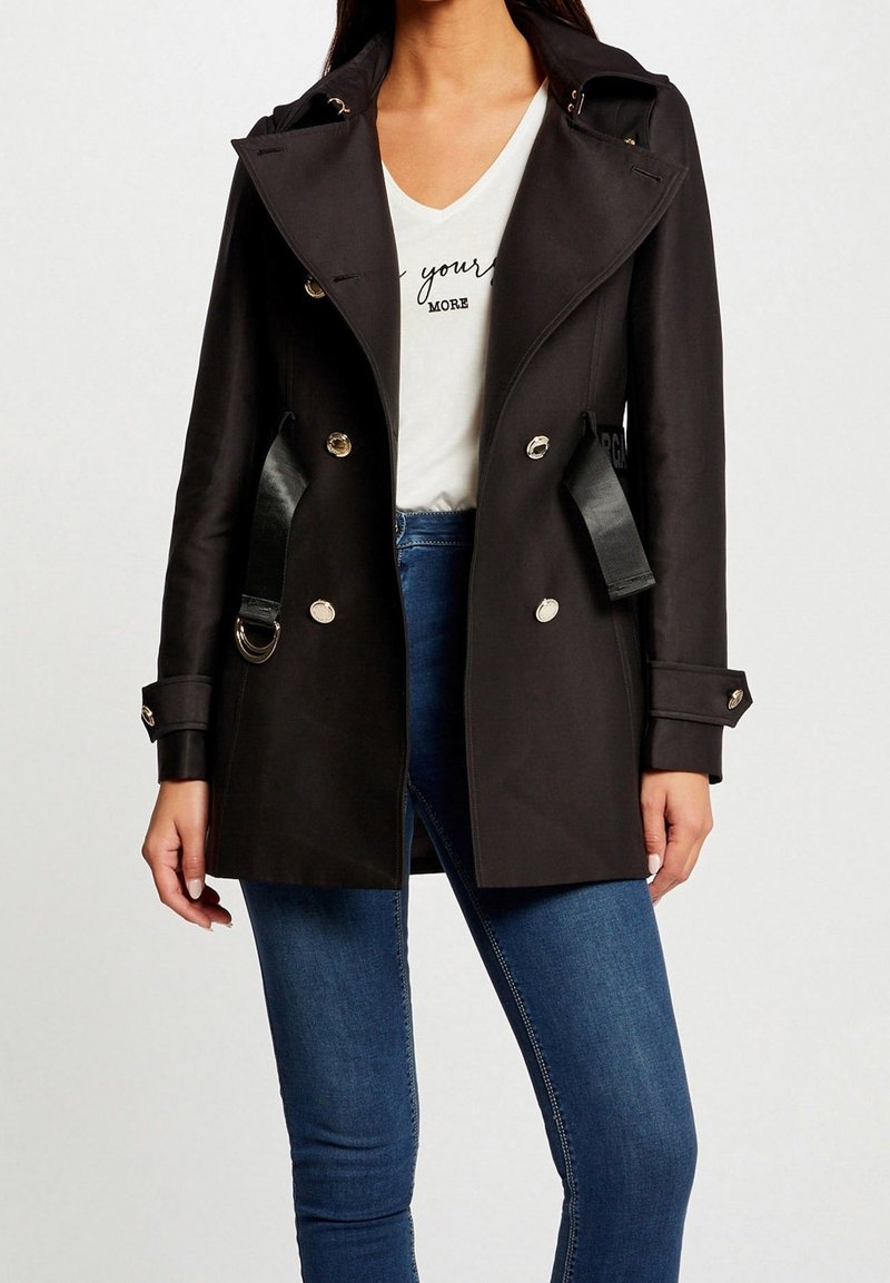 Femme portant un manteau noir croisé avec des boutons brillants sur un t-shirt blanc à col en V et un jean bleu sur un fond uni.