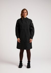 Zwarte waterdichte jas met hoge kraag, lange mouwen en een lengte tot onder de knieën. Voorzien van een badge op de schouder en een gladde textuur.