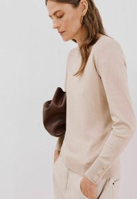 Beige gestrickter Pullover mit langen Ärmeln und gerippten Bündchen. Verfügt über drei Knopfakzente am Ärmel. Das Modell hält eine braune Tasche.