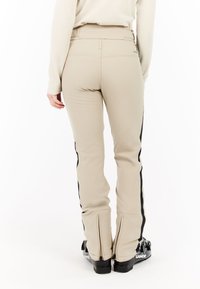 Persoon in beige skibroek met zwarte zijstrepen en zwarte skischoenen, gezien vanaf de achterkant tegen een witte achtergrond.